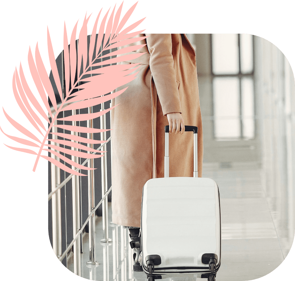 A business traveller in a long beige coat walking through an airport terminal pulling a small case
